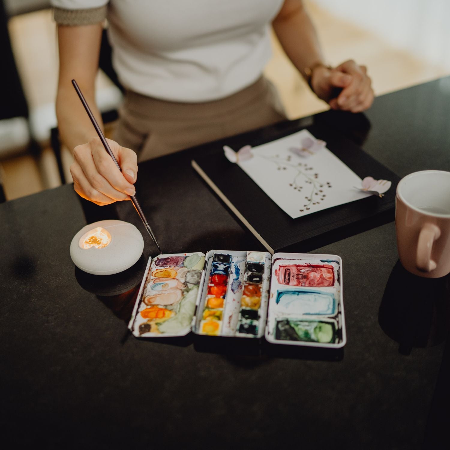 Eine Frau malt eine Grußkarte mit Aquarellfarben. Das HerzLicht liegt neben ihr und dient als Timer für ihre Kreativroutine.